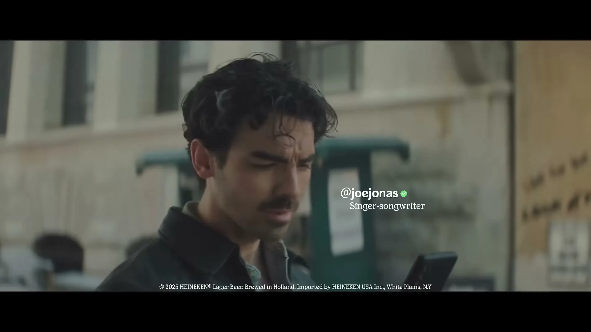 Joe Jonas, a man with dark curly hair and a mustache, looks around and then down at his phone on a city street during the day.