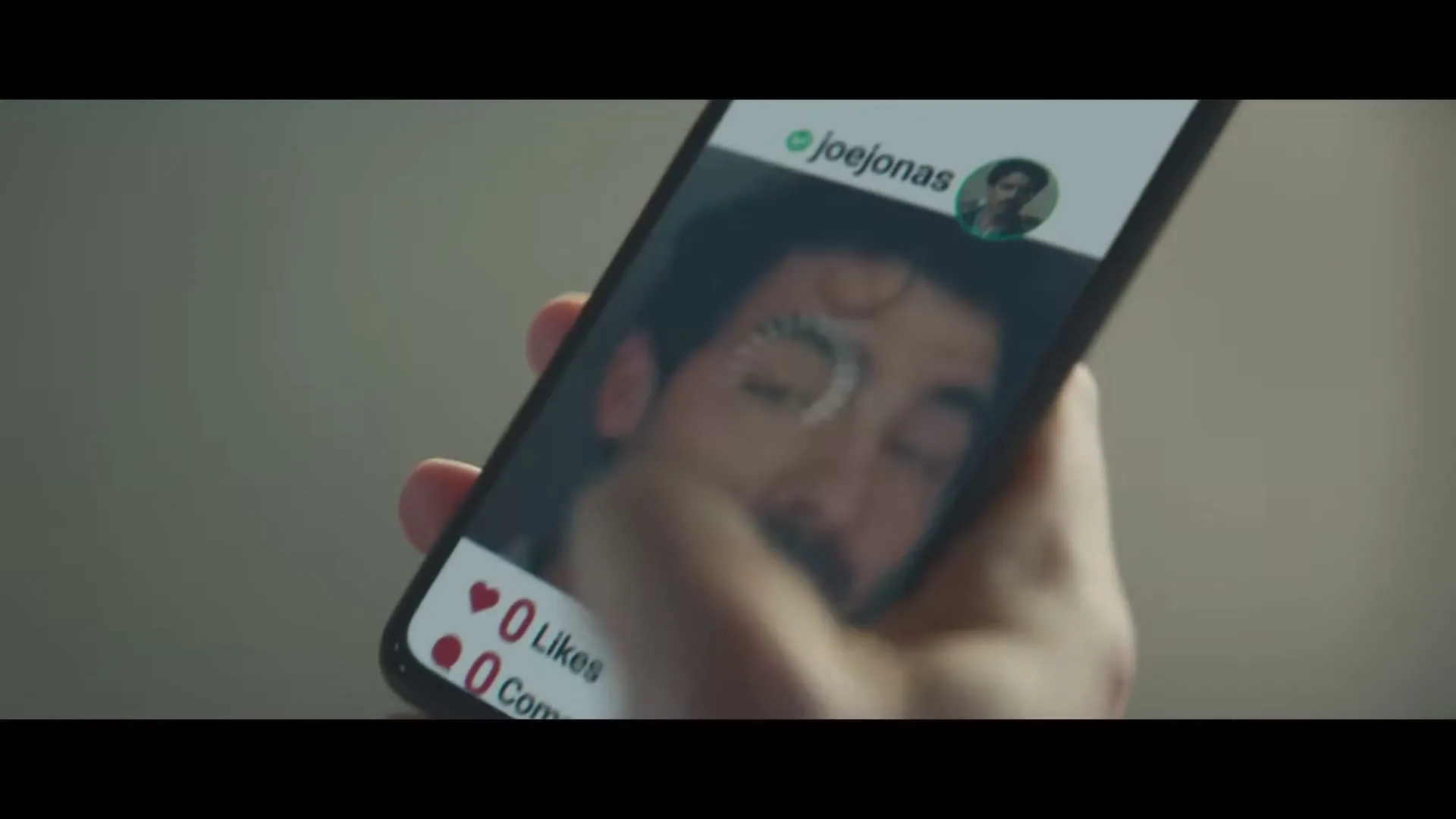 A hand holds a smartphone displaying a social media post by @joejonas, who is seen winking and smiling at the camera.