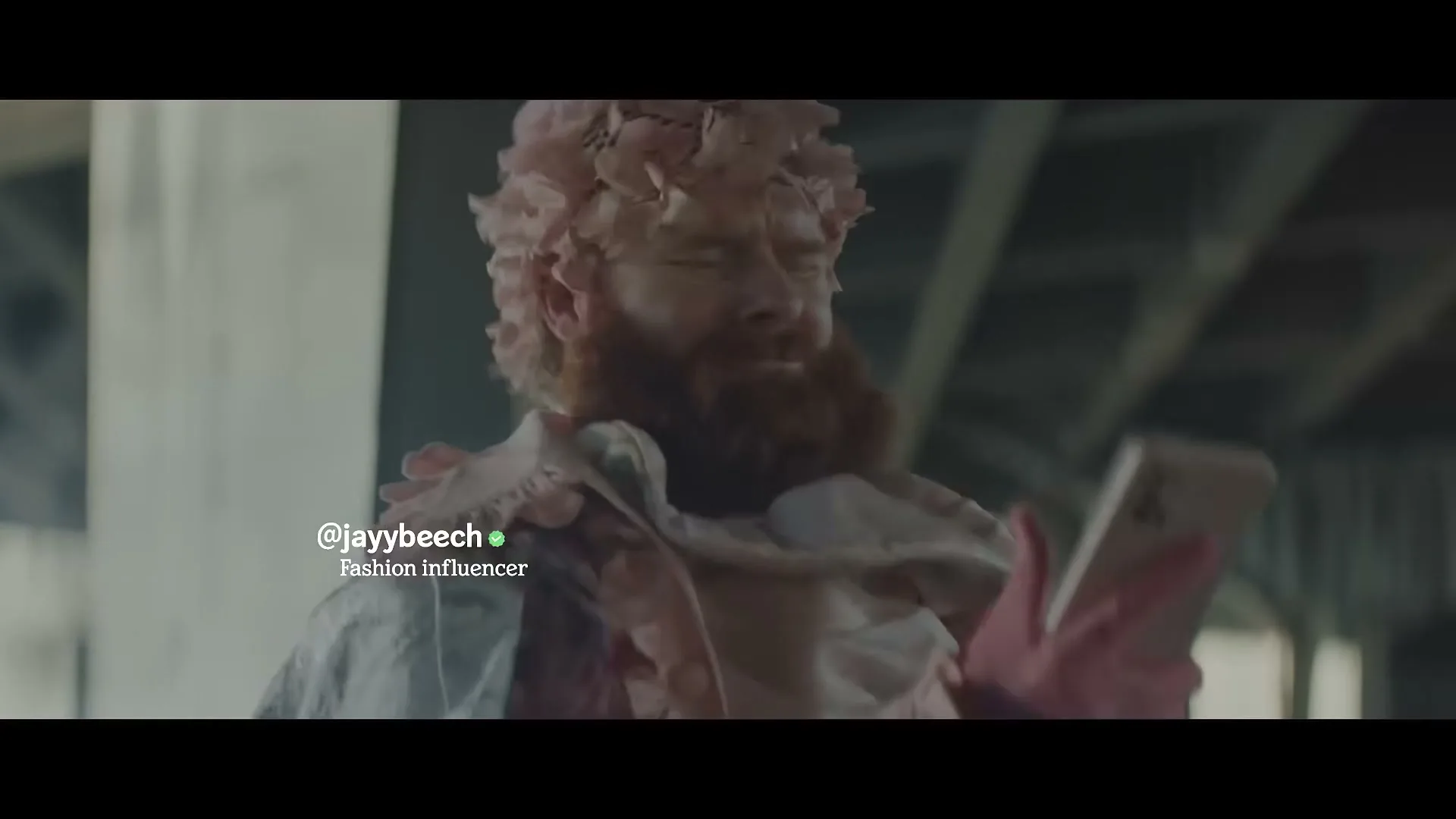 A man with a beard and pink, flower-like adornments on his head and shoulders looks at his phone under a bridge.