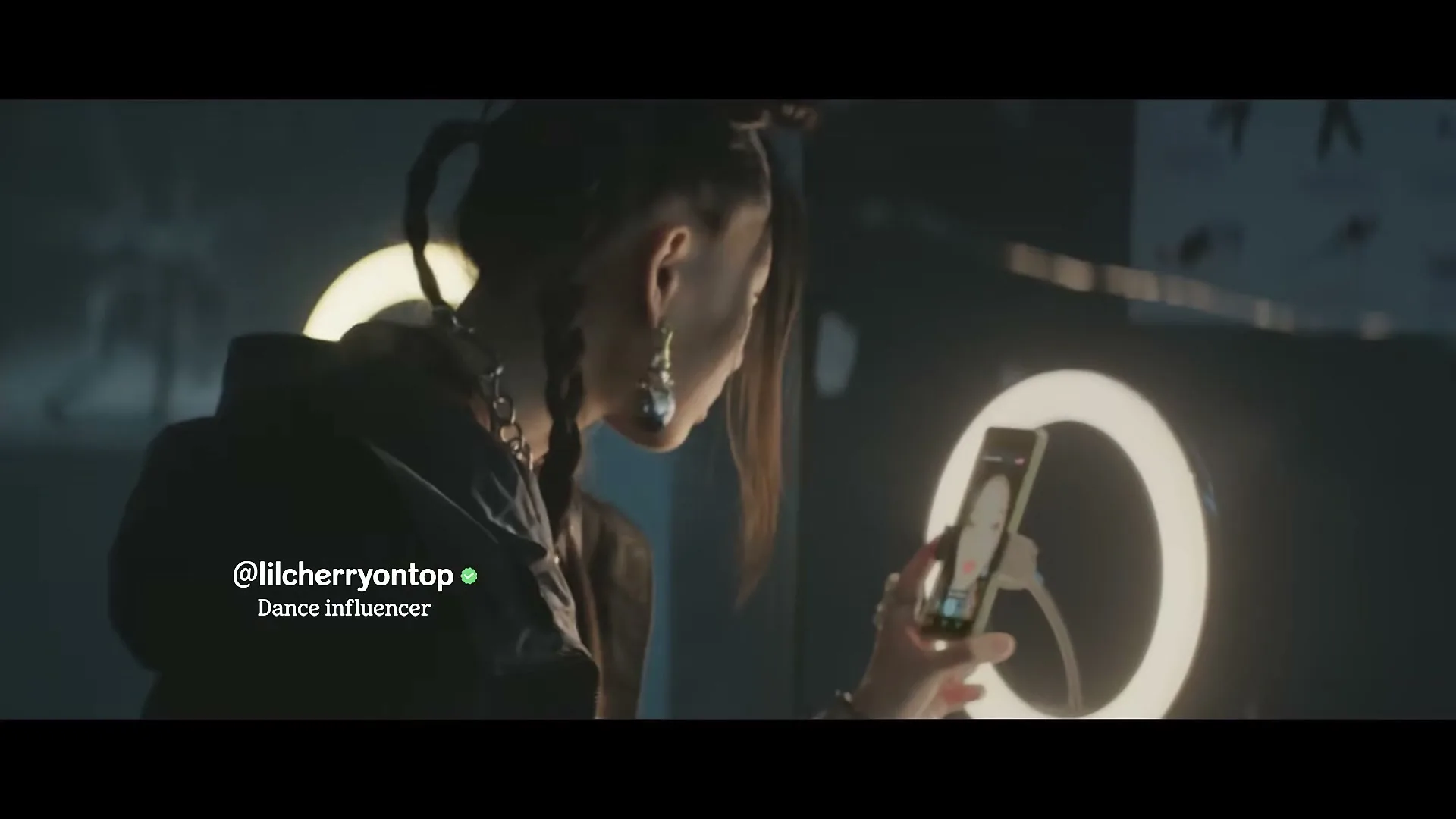 A young woman with braids and large earrings looks at her phone, which is mounted on a ring light, in a dimly lit room.
