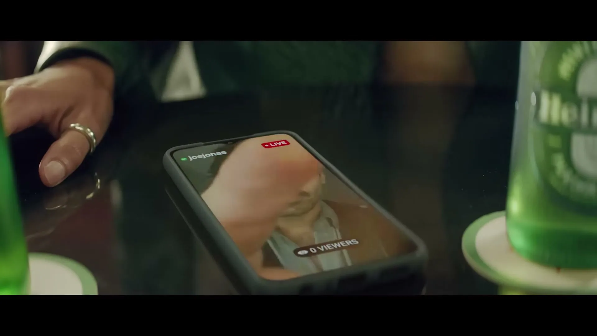 A person's hand taps a smartphone screen showing a live video of Joe Jonas with zero viewers, next to a Heineken bottle on a dark table.