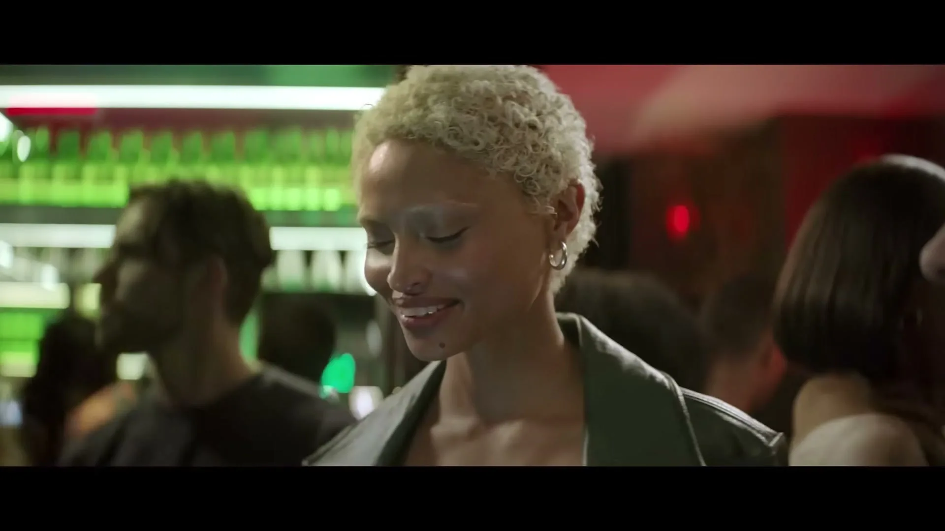 A young woman with short blonde hair and a leather jacket smiles in a crowded bar with green neon lights in the background.