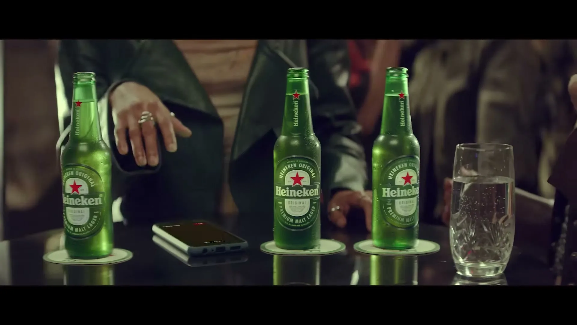 Three green Heineken beer bottles and a glass of water sit on a dark bar counter, with a person's hands visible in the background.