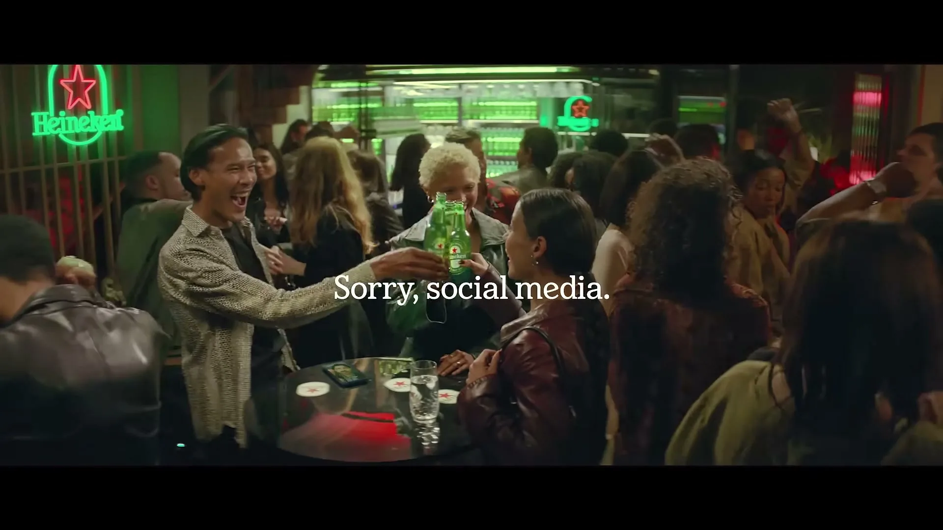 A diverse group of people are seen dancing and socializing in a lively bar, with three individuals in the foreground toasting with Heineken bottles.
