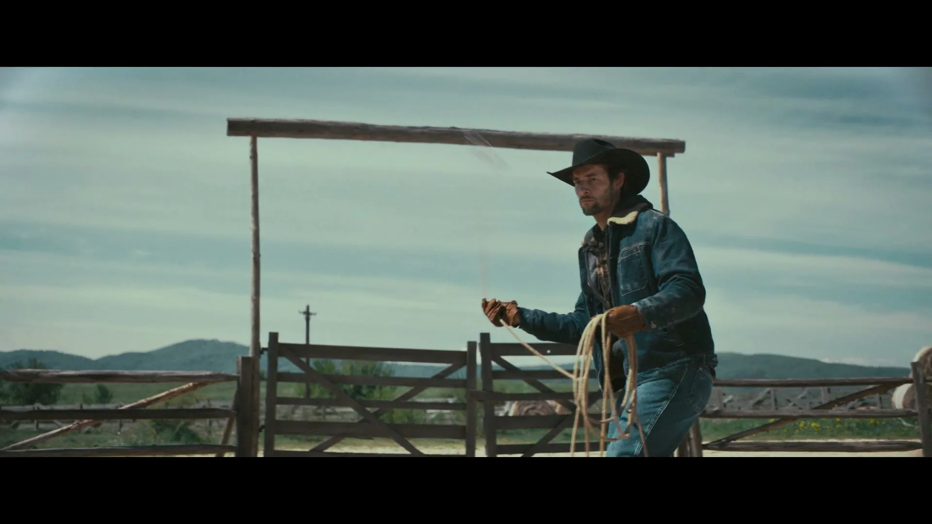 A cowboy in a denim jacket and hat practices lassoing with a rope in a dusty outdoor corral with a wooden gate and mountains in the background.