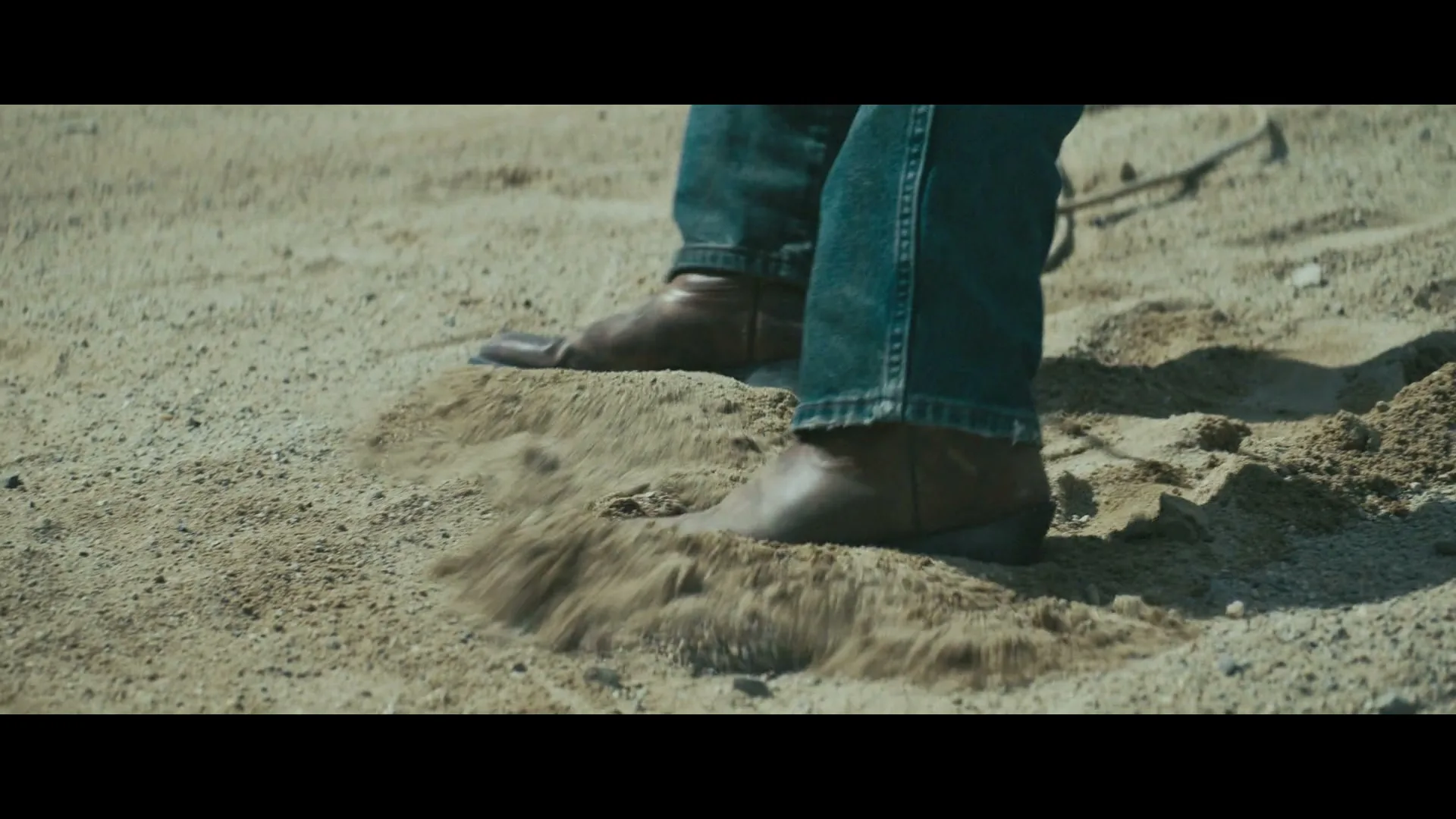 A person wearing blue jeans and brown boots kicks up sand on a sunny day, creating a small cloud of dust.