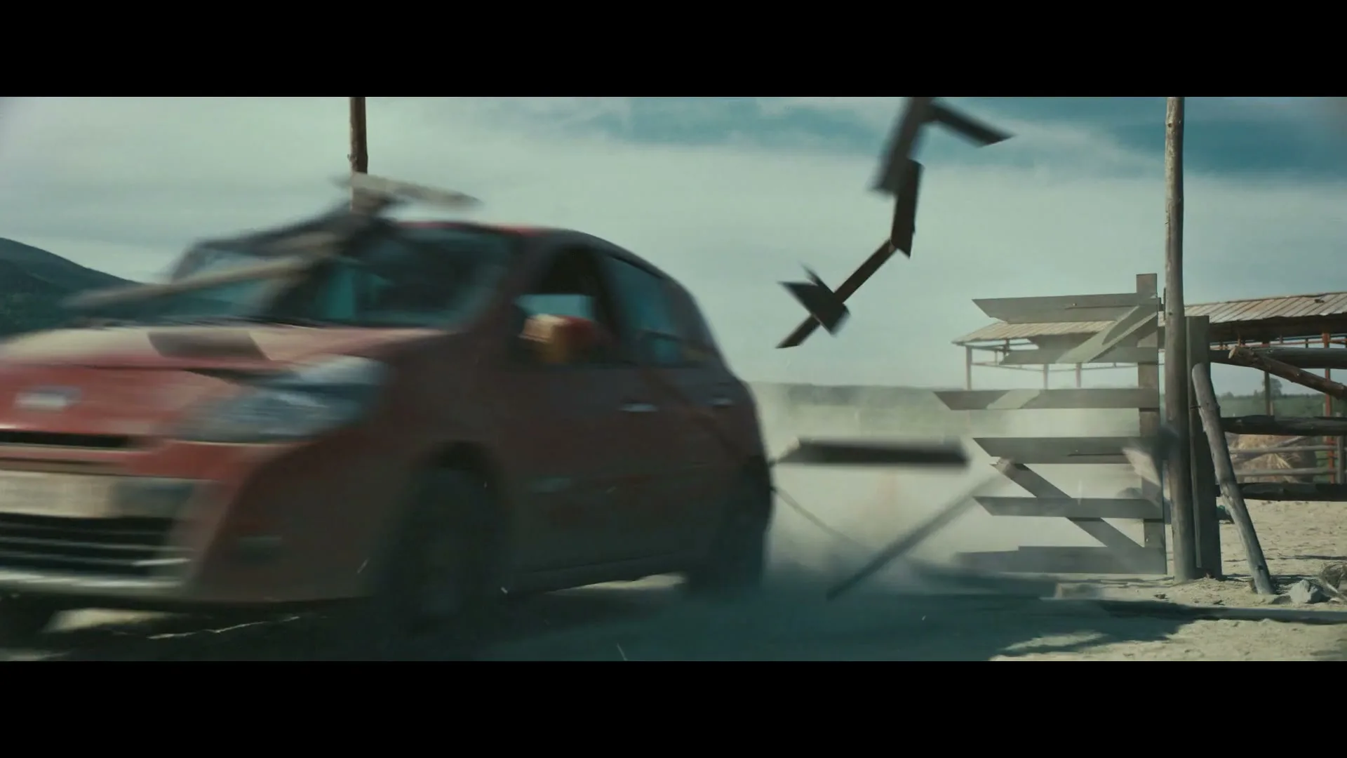 A red car crashes through a wooden fence gate, kicking up a cloud of dust in a rural, mountainous landscape under a cloudy sky.