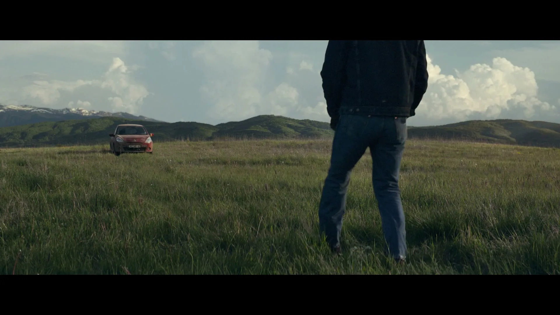A man in a cowboy hat and denim jacket walks towards a red car in a vast green field with mountains in the background, then kneels.