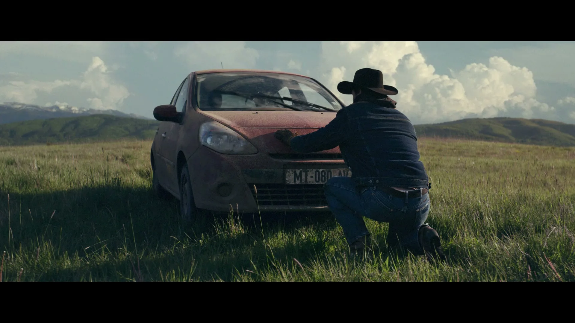 A man in a cowboy hat kneels in a grassy field, wiping dust off the hood of a red car under a cloudy sky.
