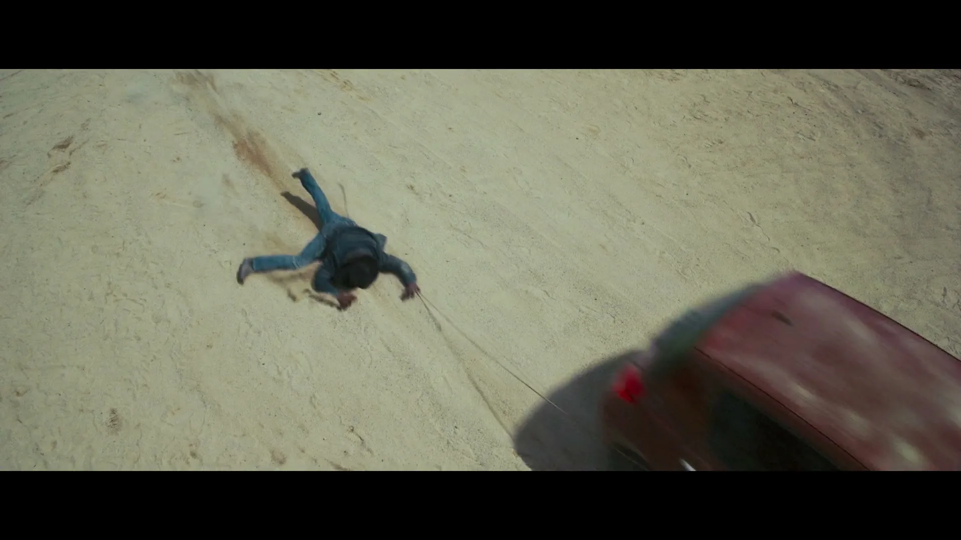 A man in blue jeans and a dark jacket is dragged across a sandy, dusty terrain by a red car, seen from an overhead perspective.
