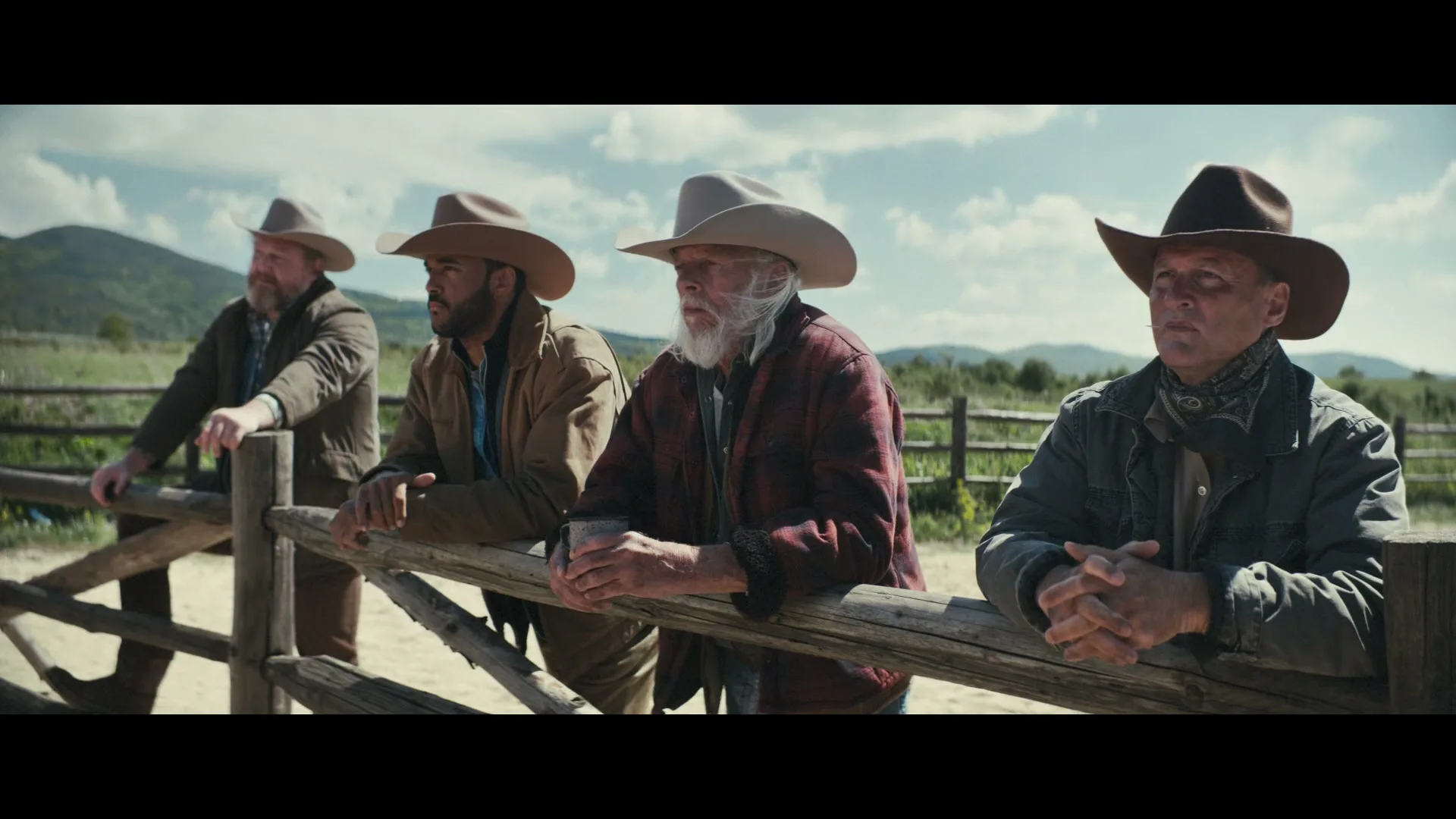Four male cowboys, dressed in western attire, lean on a wooden fence, looking off-camera in a rural, mountainous landscape.