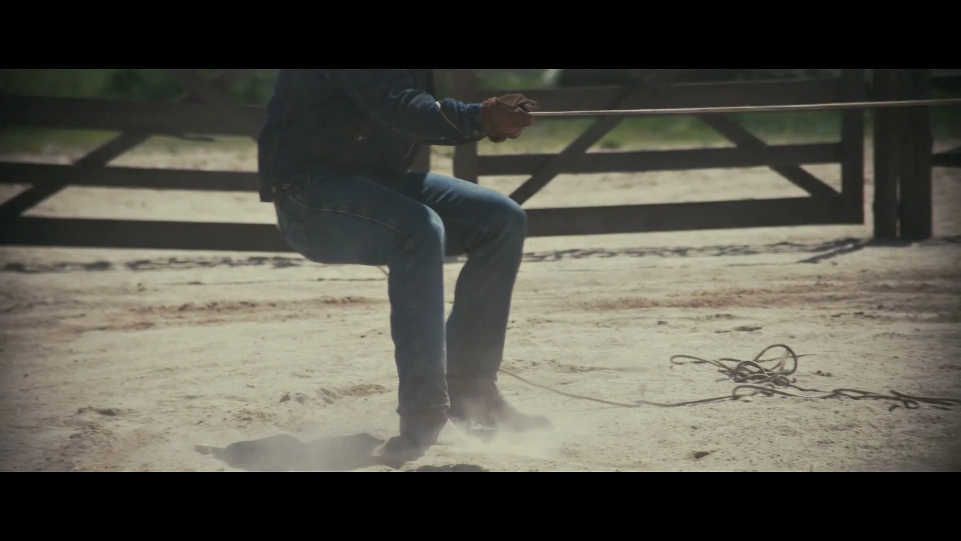 A person in jeans and a denim jacket pulls a rope, kicking up dust in a sandy outdoor area with a wooden fence.