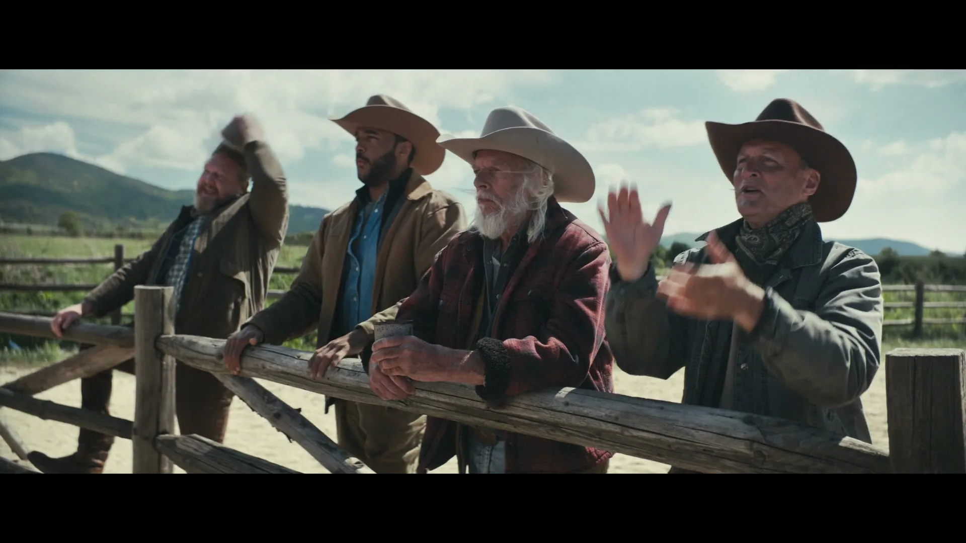 Four cowboys, dressed in rugged western wear, lean on a wooden fence, watching something off-screen with expressions of excitement and applause on a sunny day.