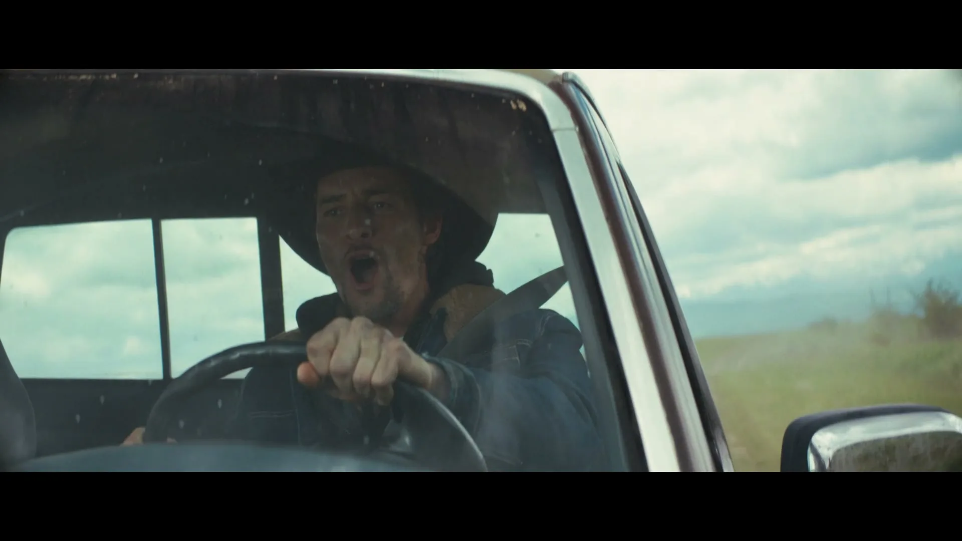 A man in a cowboy hat and denim jacket drives a pickup truck on a dirt road, yelling with excitement.