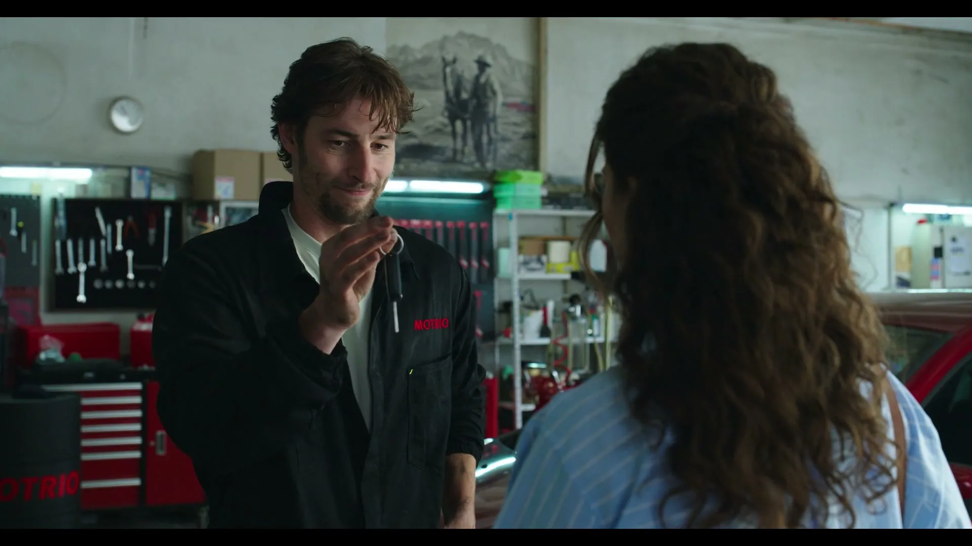 A male mechanic in a black jumpsuit hands a car key to a female customer with long curly hair in a garage.