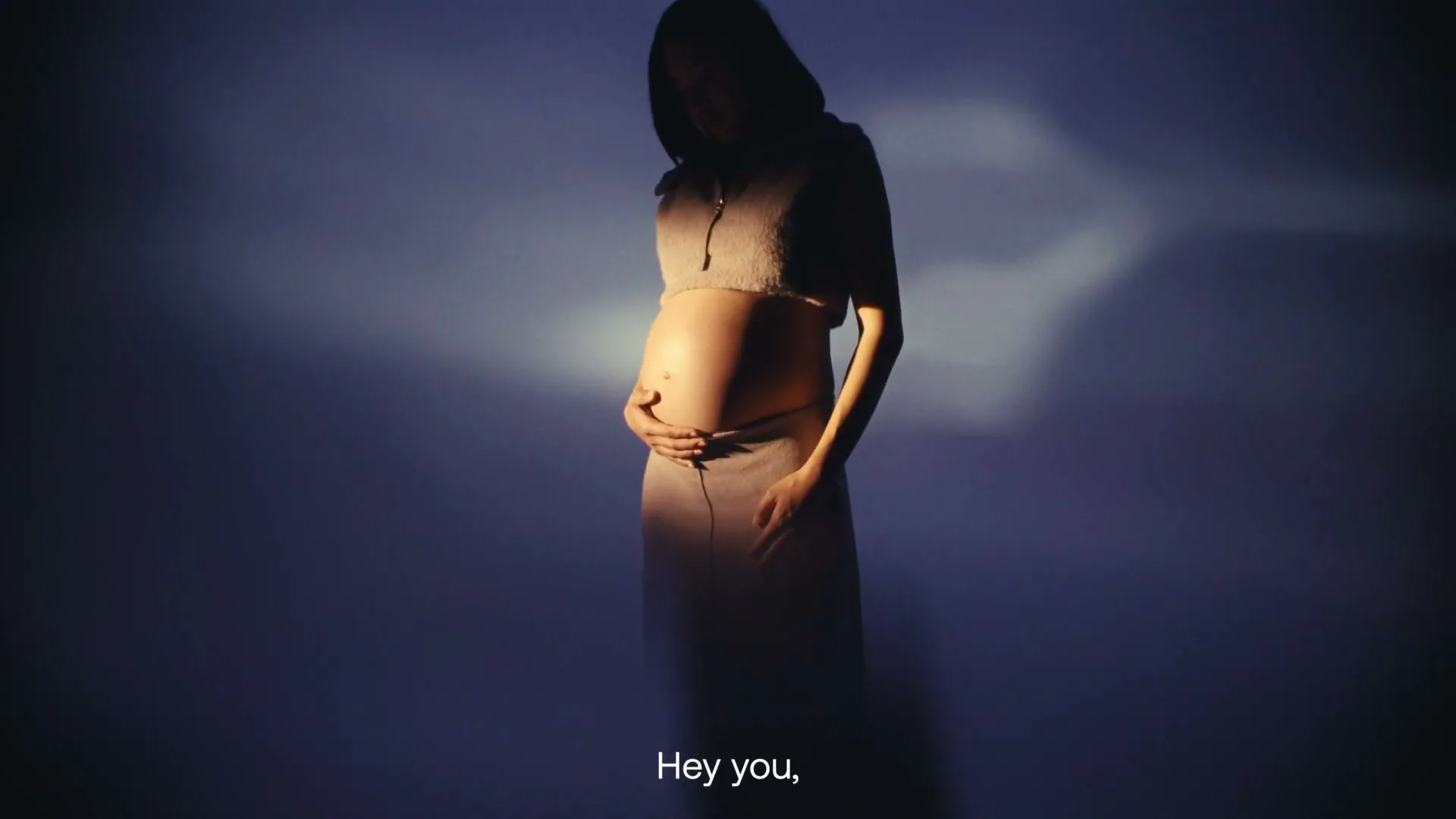 A pregnant woman in a cropped top and skirt stands in a spotlight, gently cradling her baby bump in a studio.