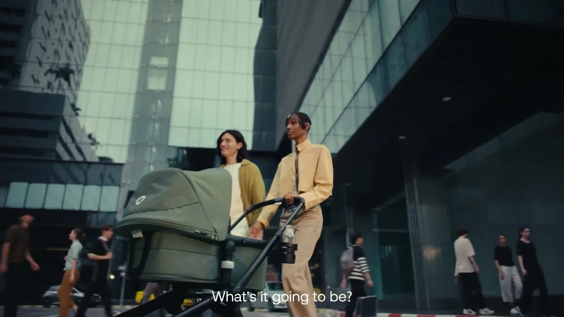 Two young adults, one male and one female, push a green Bugaboo stroller down a city street with modern buildings in the background.