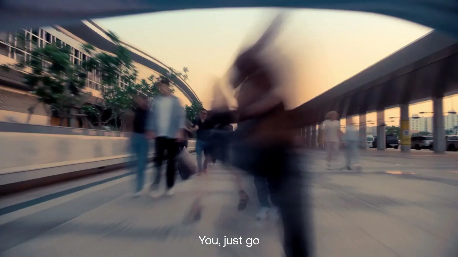 A low-angle shot shows a crowd of people walking quickly on a wide outdoor walkway at sunset, with motion blur emphasizing their movement.