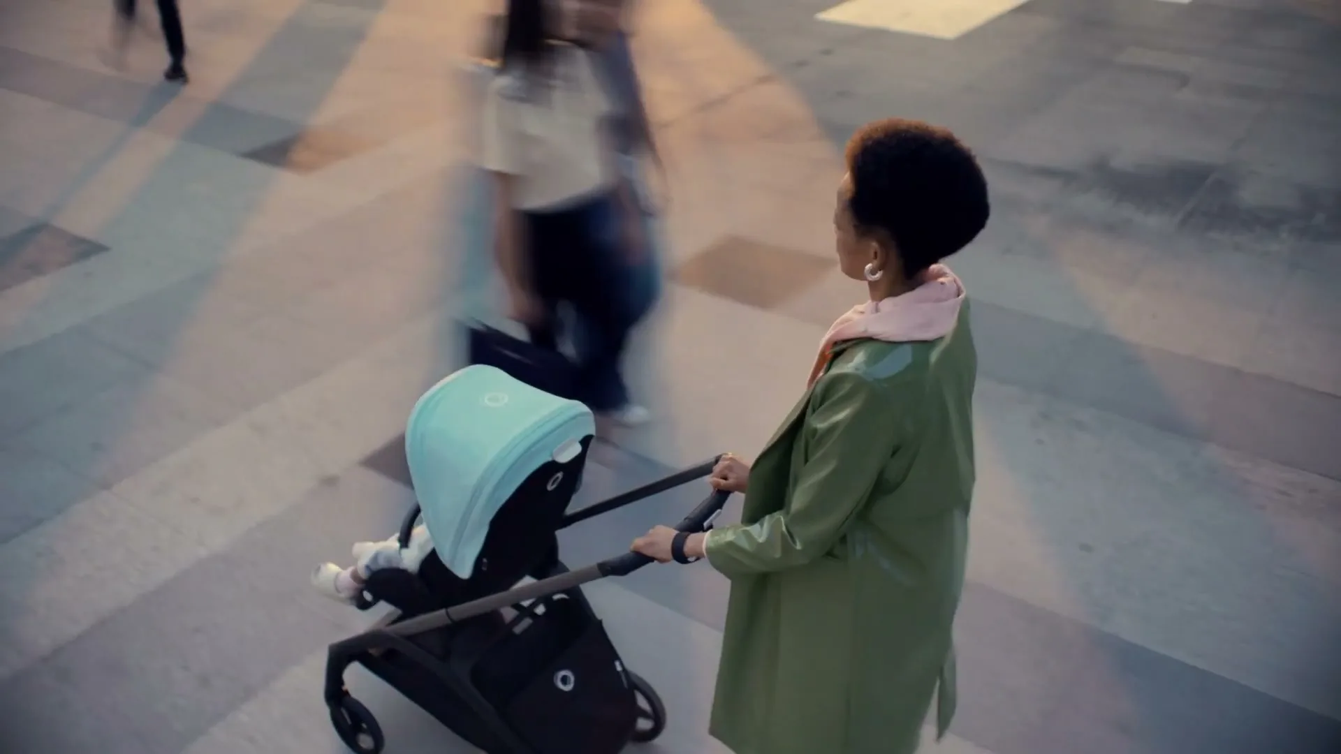 A Black woman in a green coat pushes a baby stroller with a light blue canopy through a busy urban square at dusk.