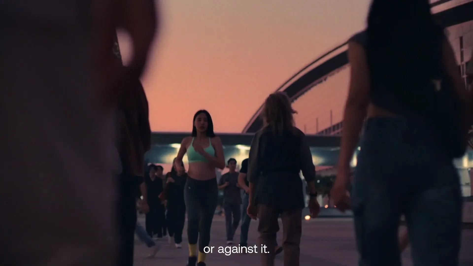 A diverse group of people, some in athletic wear, walk and run towards the camera on a city street at dusk, with a bridge in the background.