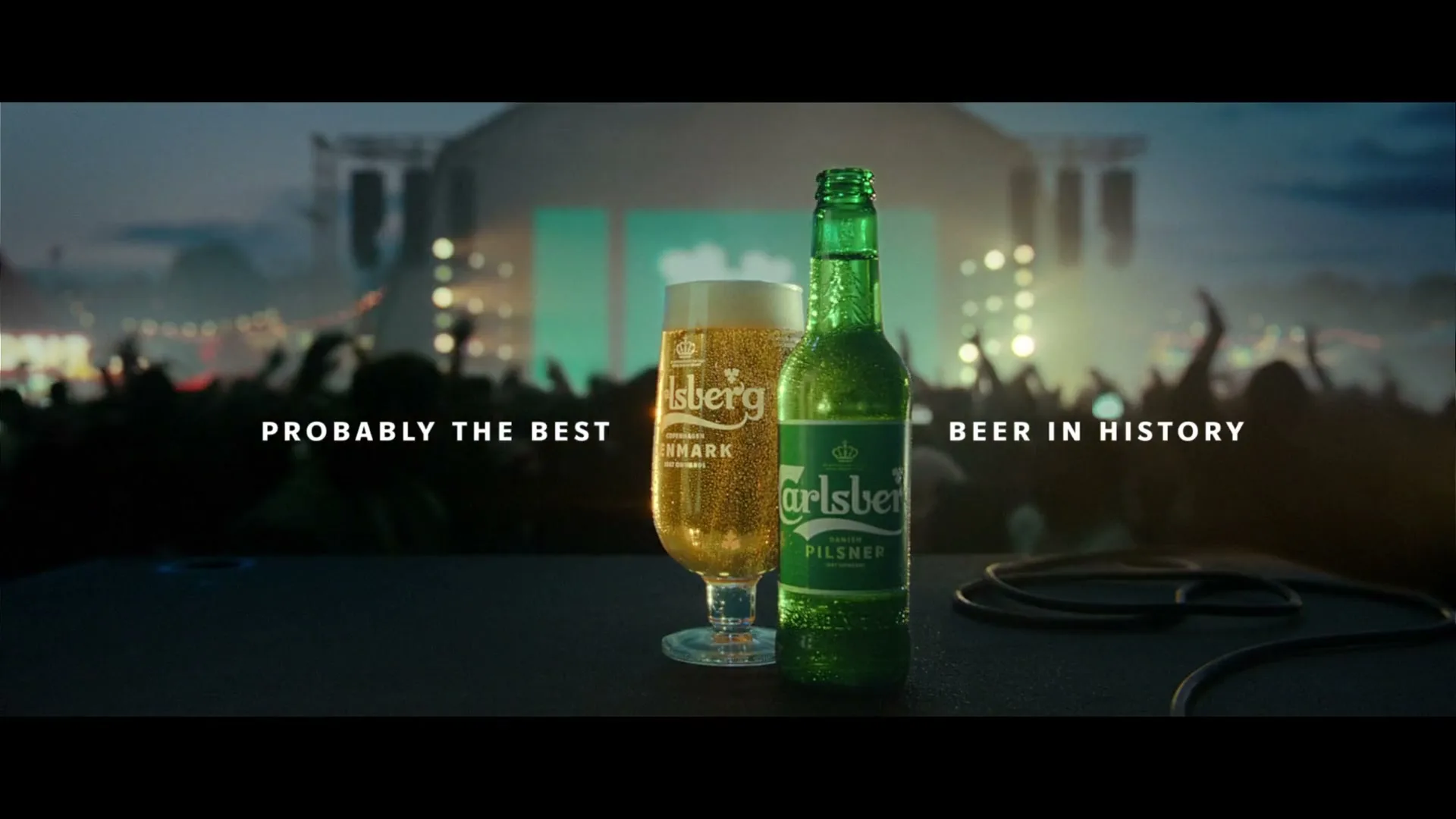 A Carlsberg beer bottle and a glass of beer sit on a stage, with a blurred crowd and concert lights in the background.