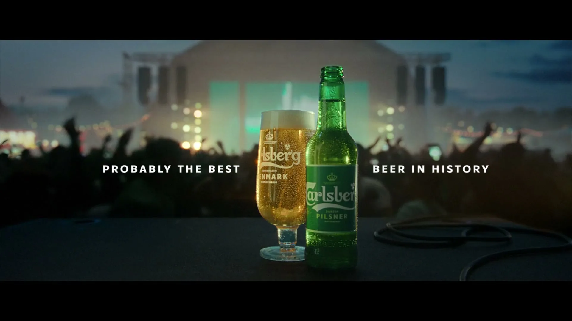 A Carlsberg beer bottle and glass sit on a stage with a blurred concert crowd in the background, featuring the tagline "PROBABLY THE BEST BEER IN HISTORY."