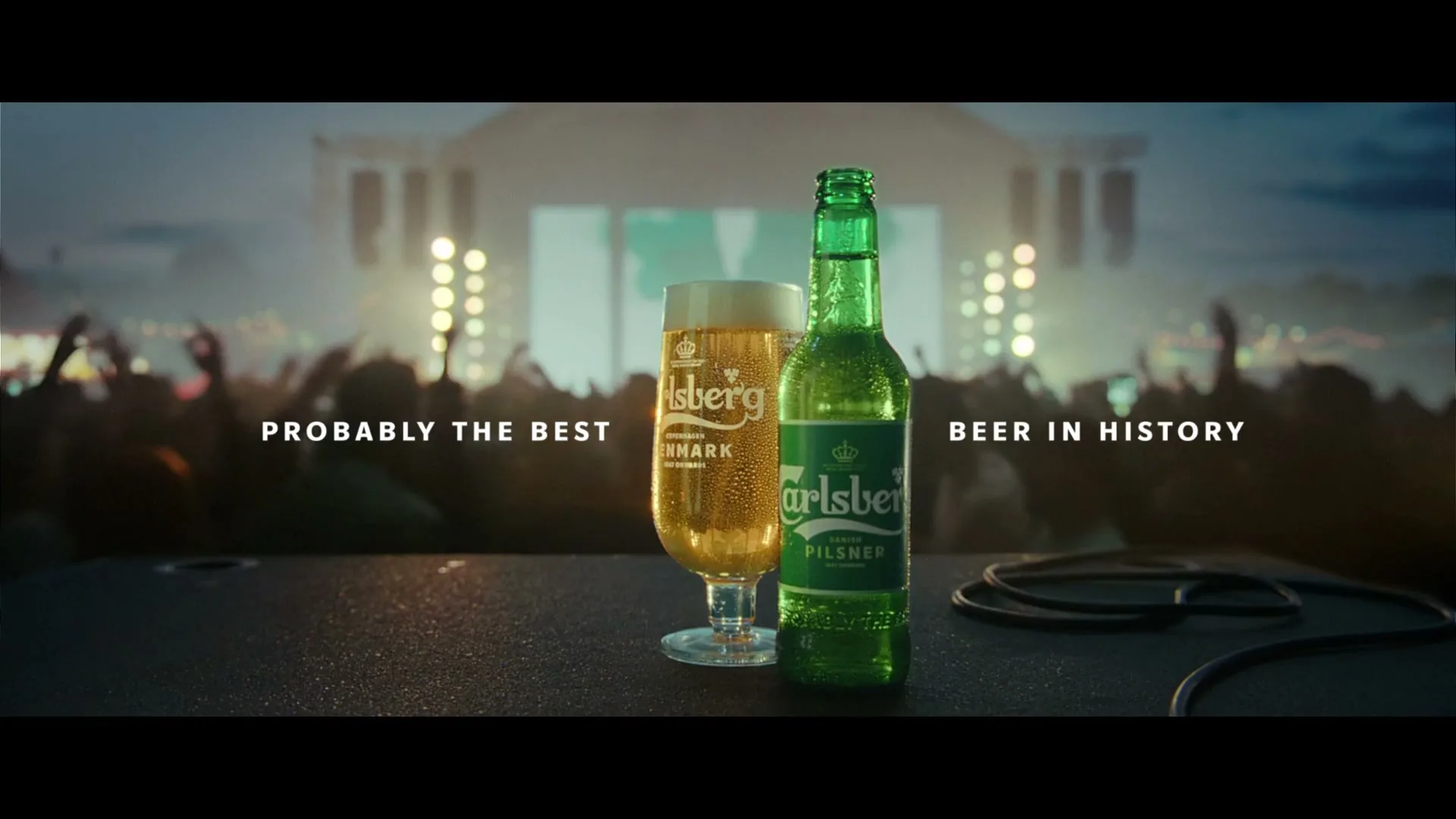A Carlsberg beer bottle and a glass of beer sit on a stage, with a blurred concert crowd and stage lights in the background.
