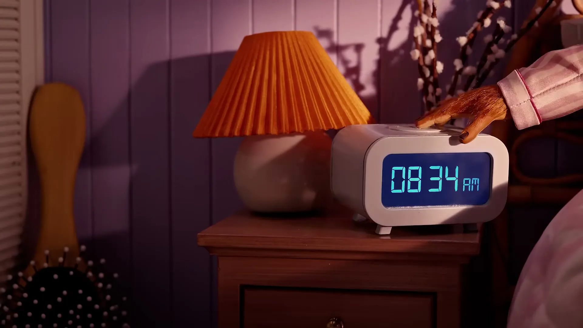 A monkey's hand in pink striped pajamas reaches out to turn off a white digital alarm clock on a wooden nightstand next to a lamp.
