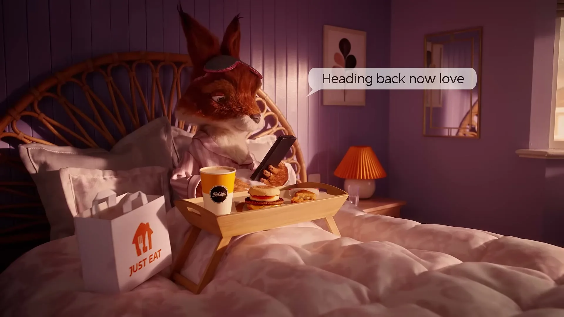 A fox character in pajamas sits up in bed, looking at a phone while a breakfast tray and a Just Eat bag are on the bed.