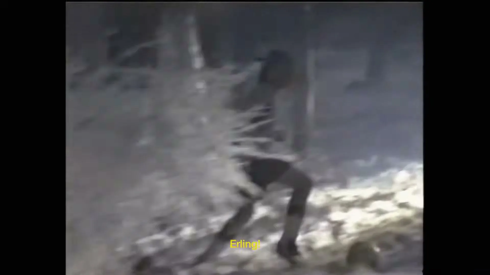 A young boy in a t-shirt and shorts runs through a snowy forest, kicking up snow as he moves, captured in a grainy, home video style.