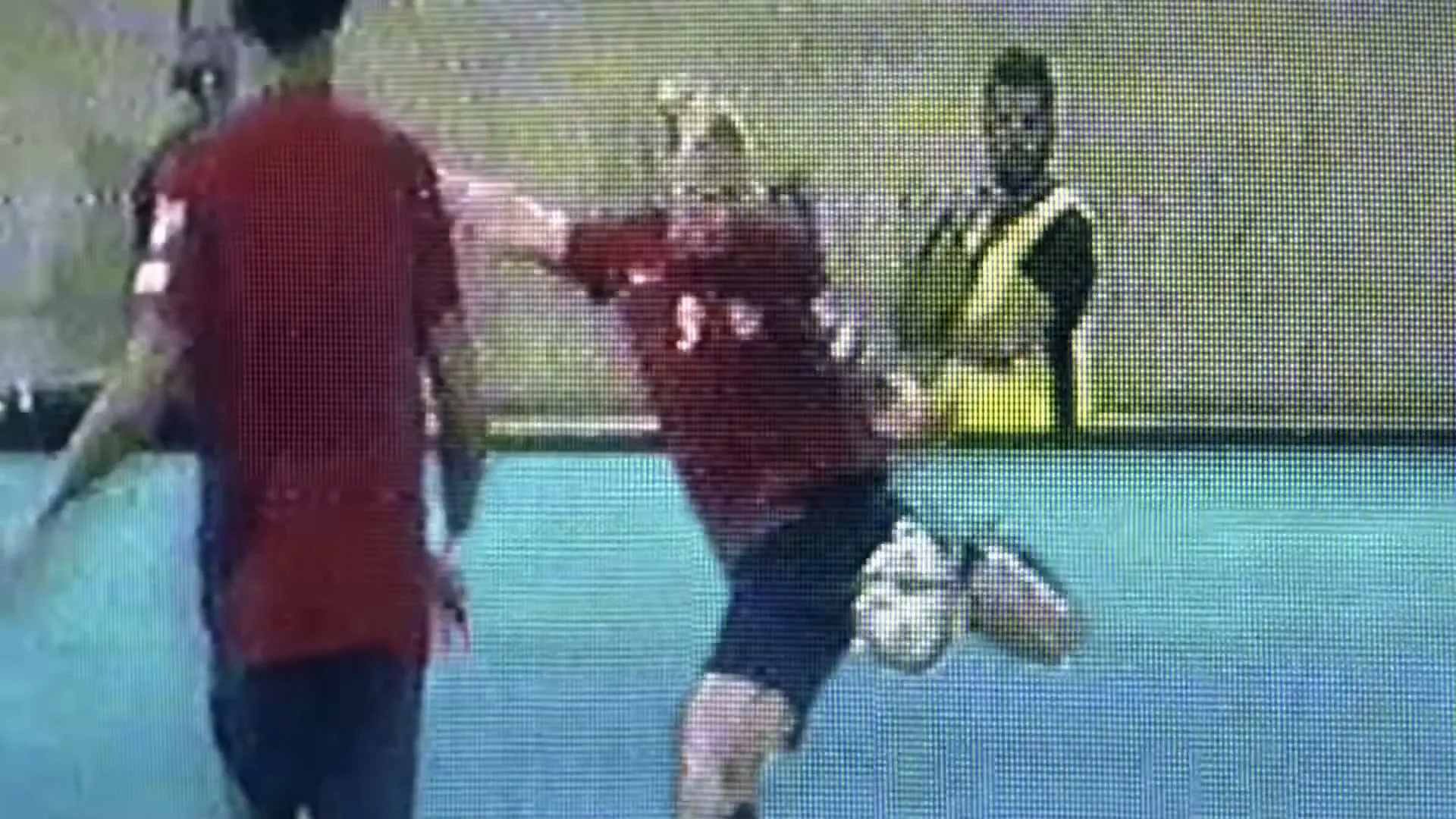 A male soccer player in a red jersey kicks a soccer ball during a game, captured by a low-quality security camera.