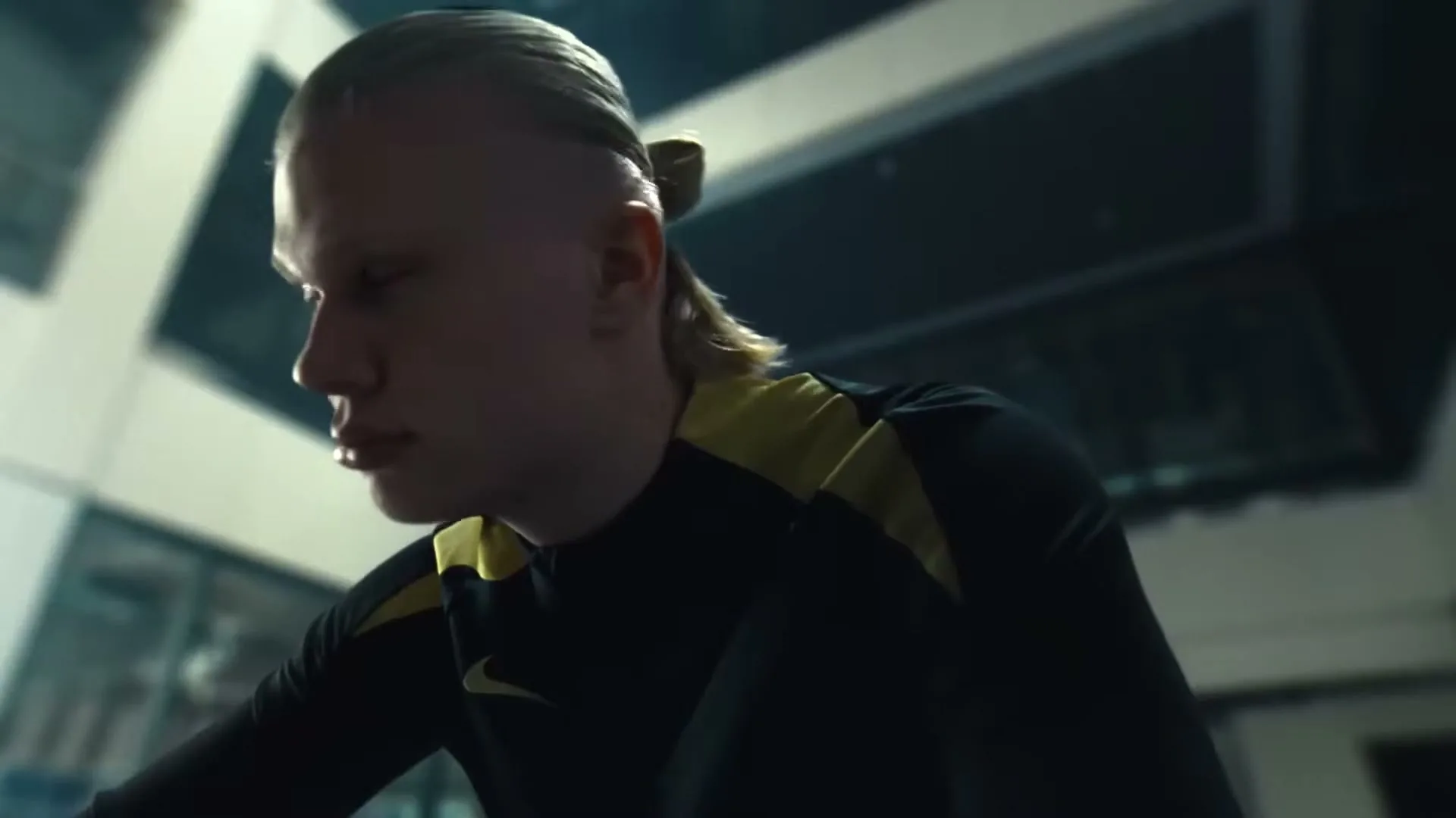 A young male athlete with a distinctive hairstyle, wearing a black and yellow athletic shirt, looks down and then turns his head to the side in an indoor setting.