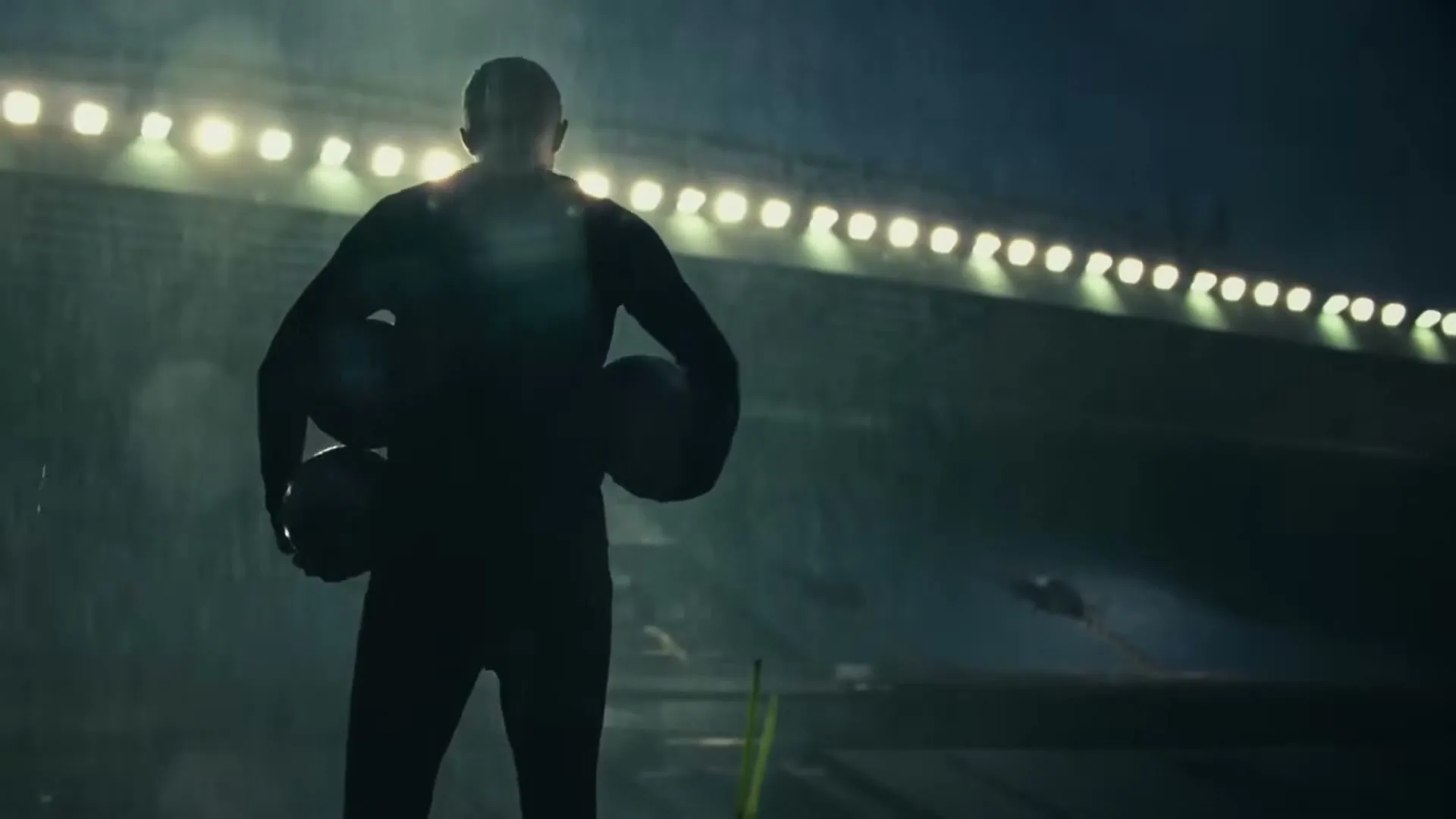 A male athlete stands silhouetted on a rainy soccer field at night, holding two soccer balls, facing away from the camera towards stadium lights.