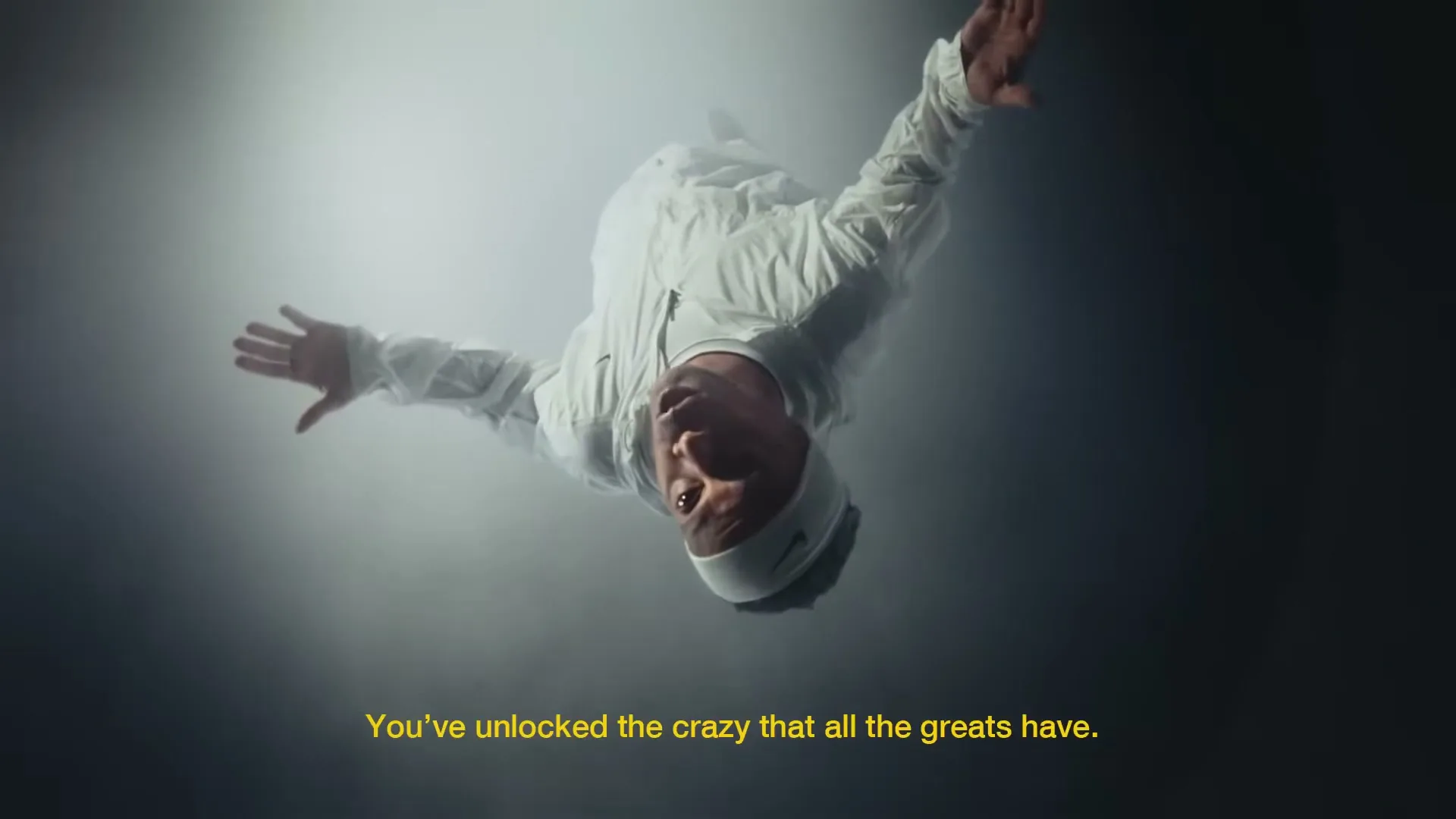 A male athlete in white sportswear and a headband floats upside down in a smoky, spotlighted void, arms outstretched.