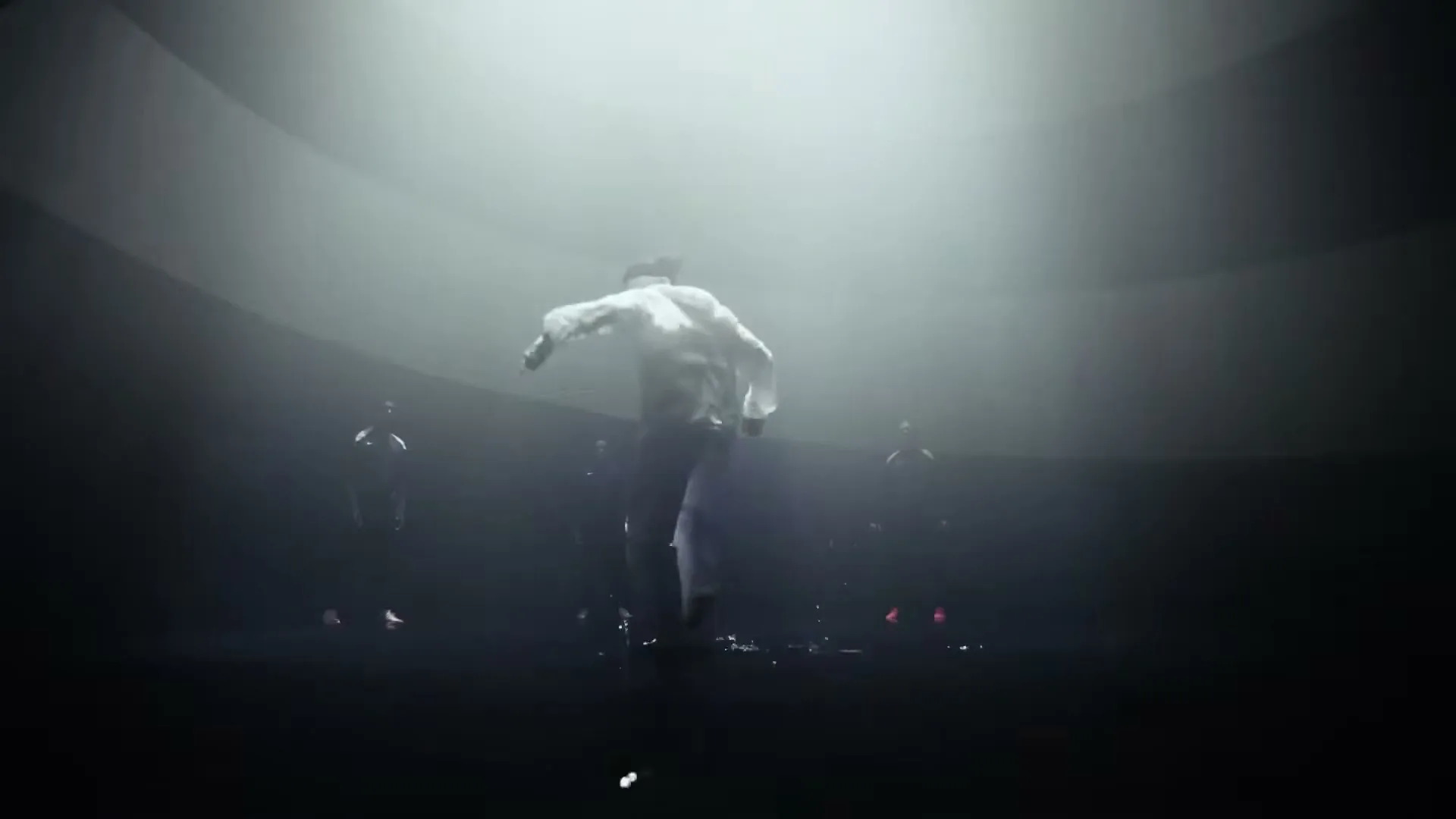 A male dancer in white clothing leaps in a spotlight, surrounded by other shadowy figures in a dark, circular room.