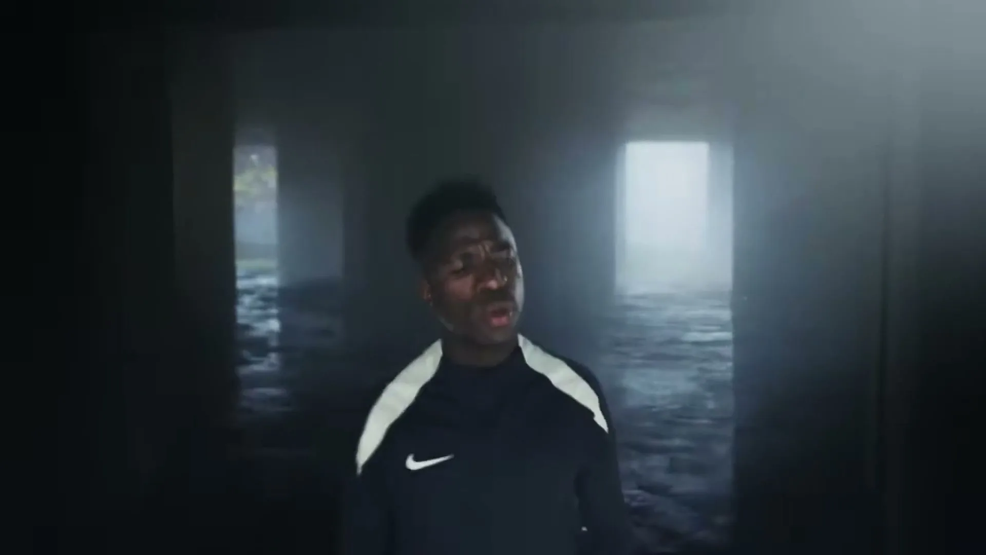 A young Black man in a dark athletic top with a white stripe and a Nike logo stands in a dark, watery tunnel, looking distressed.