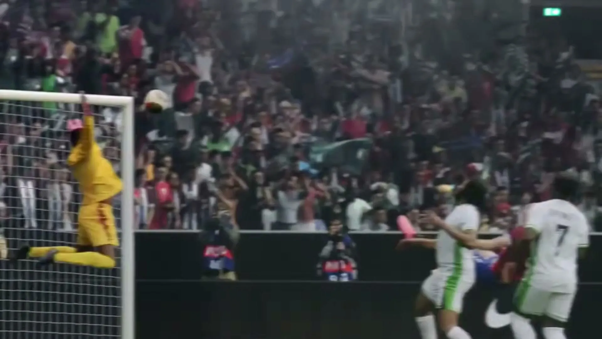 A female soccer player in a red and blue uniform kicks a soccer ball mid-air, surrounded by other players and a cheering crowd in a stadium.