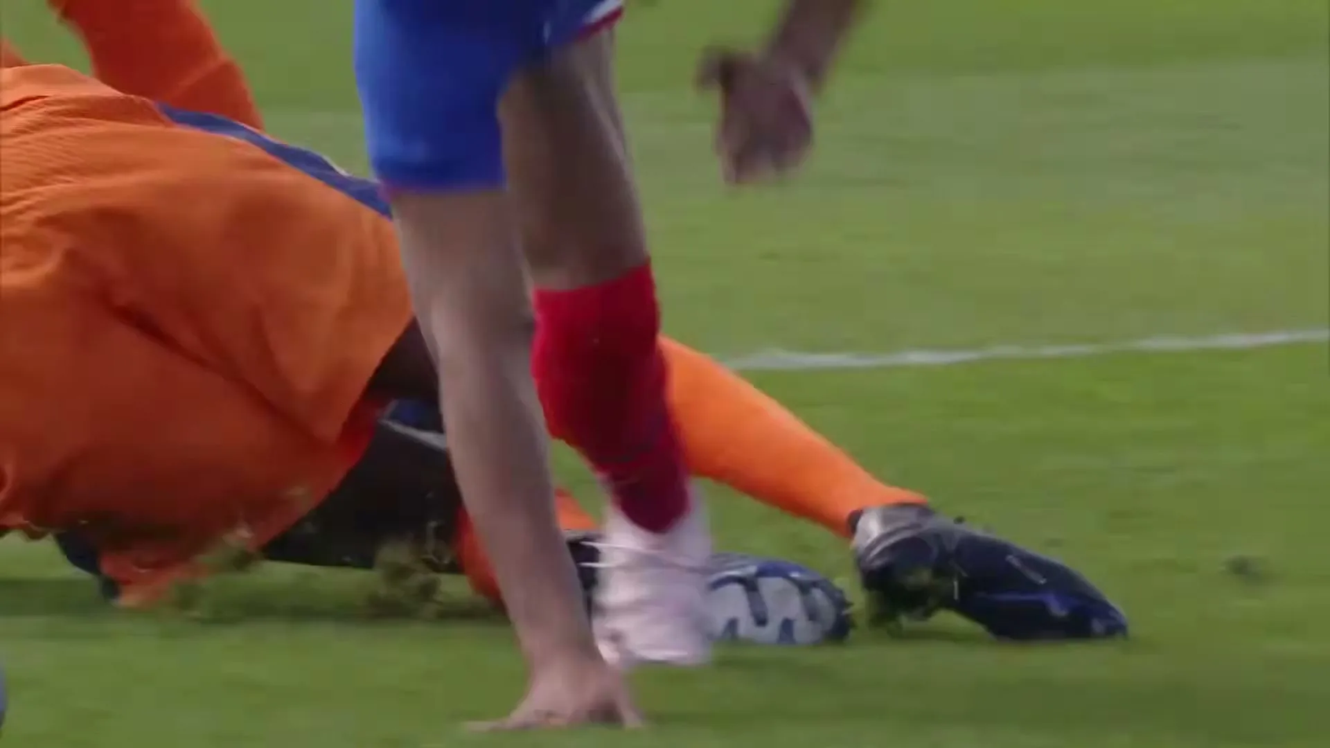 A soccer player in a blue jersey dribbles the ball, then falls to the ground after being tackled by an opponent in an orange jersey, on a green field in a stadium.