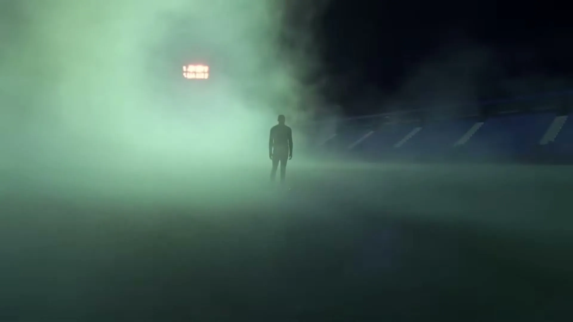 A lone male figure stands silhouetted in a stadium, surrounded by green mist, with a scoreboard visible in the distance.