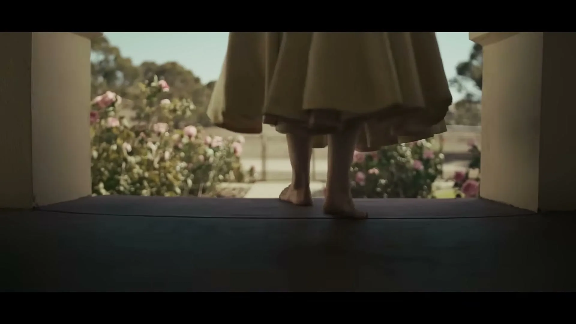 A barefoot woman in a long dress walks out of a house onto a porch, with a sunny, rose-filled garden and a dirt road in the background.