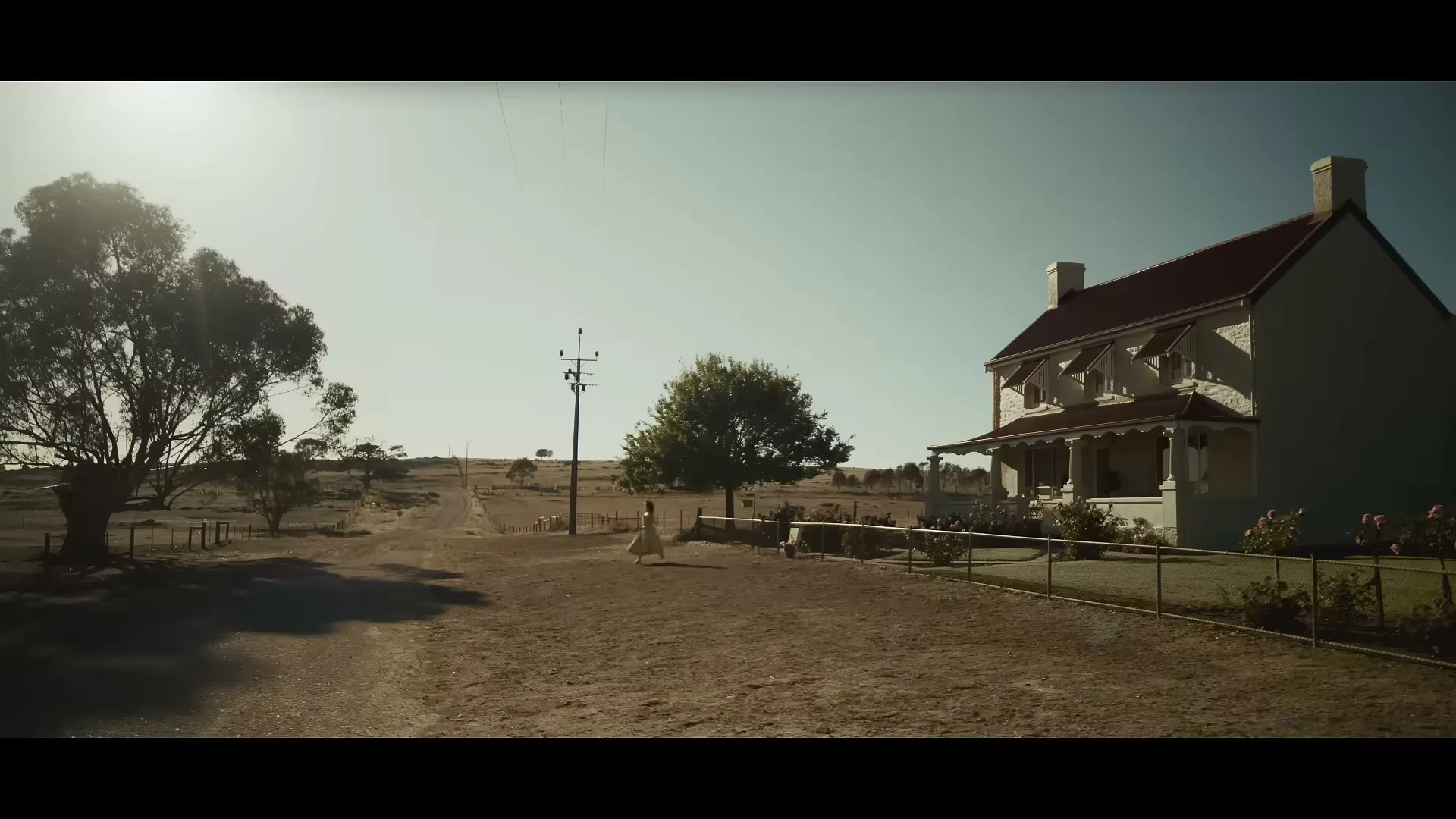A young woman in a yellow dress walks away from a large house in a rural, arid landscape under a bright sky.