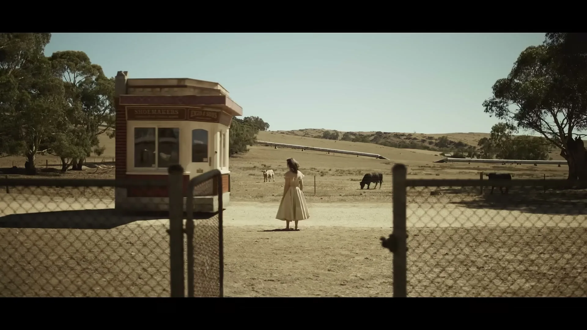 A woman in a white dress stands in a dry, rural landscape with a small building and cattle, looking towards distant hills under a clear sky.