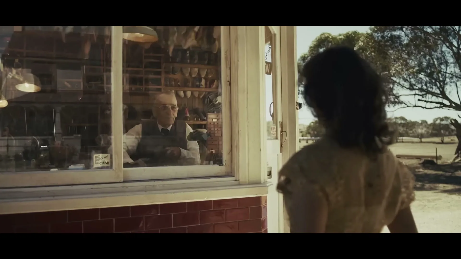 A woman with dark hair stands with her back to the camera, looking into a shop window where an older man in a vest is working, in a rural setting.