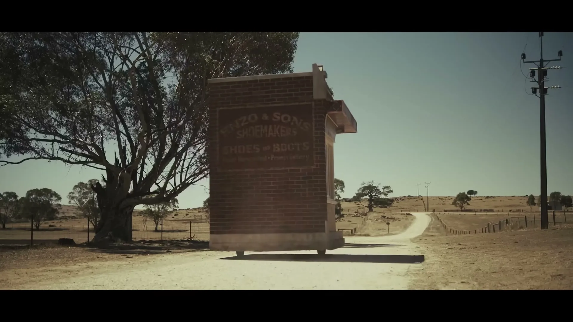 A brick building with a sign for "Enzo & Sons Shoemakers" is seen on a dirt road in a dry, rural landscape under a clear sky.