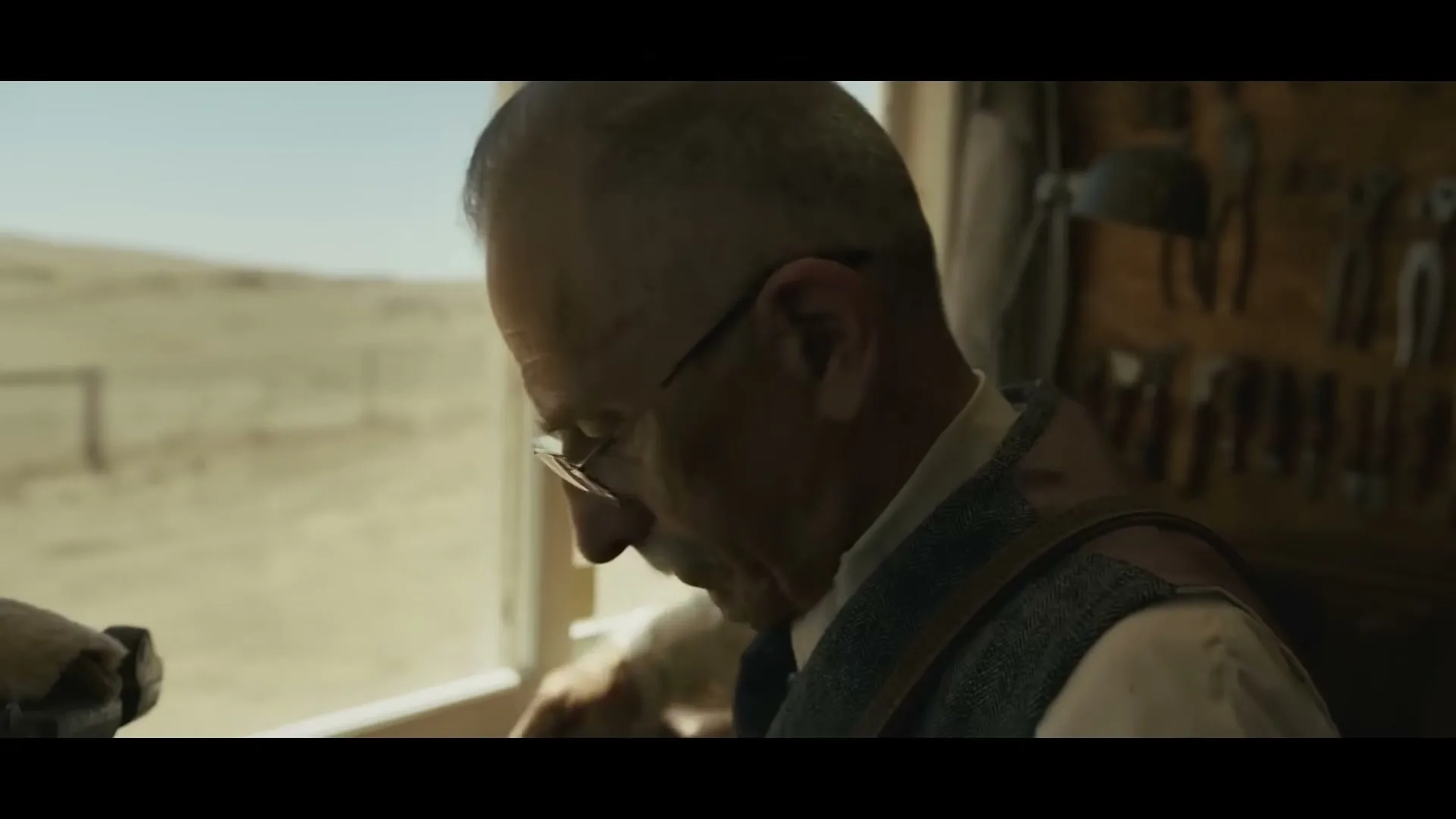 An older man with glasses and a vest works on a shoe at a workbench, with a desert landscape visible outside a window.