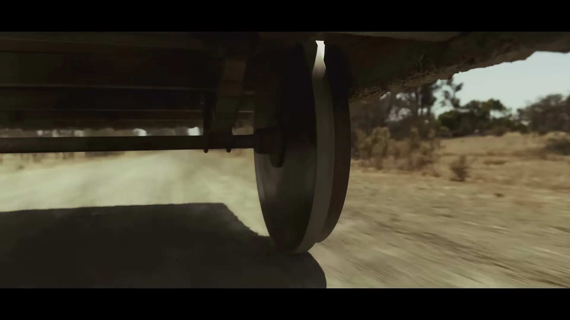 A low-angle shot shows a wagon wheel kicking up dust on a dirt road in a dry, rural landscape.
