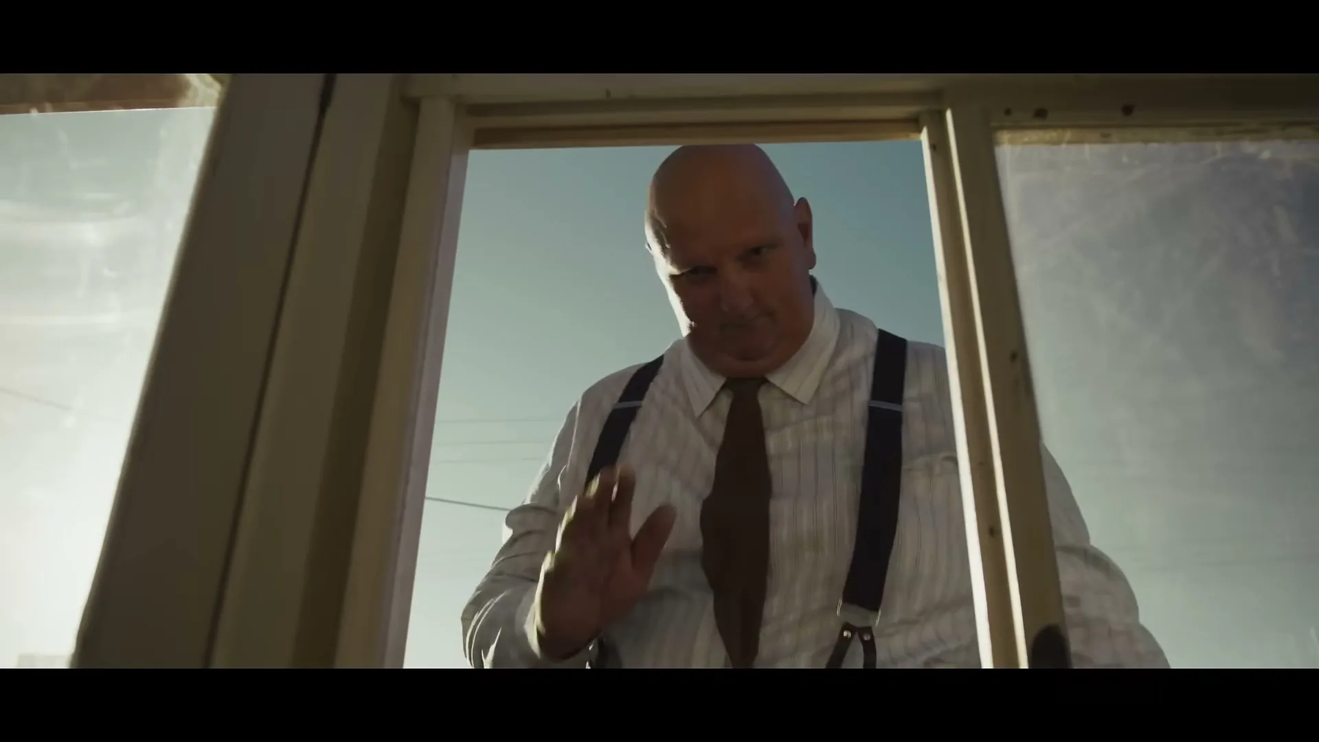 A bald man in a white shirt, brown tie, and suspenders looks down and waves from outside a window, with a bright sky in the background.