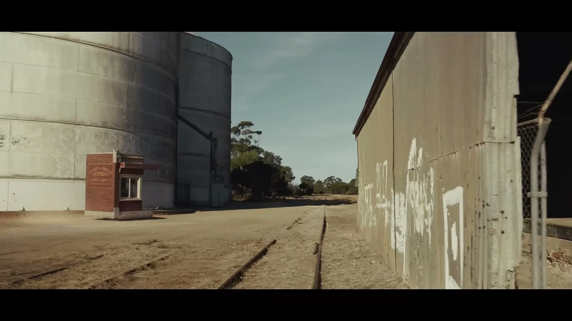 A wide shot of an abandoned train track with a large silo and a corrugated iron building on either side under a clear sky.