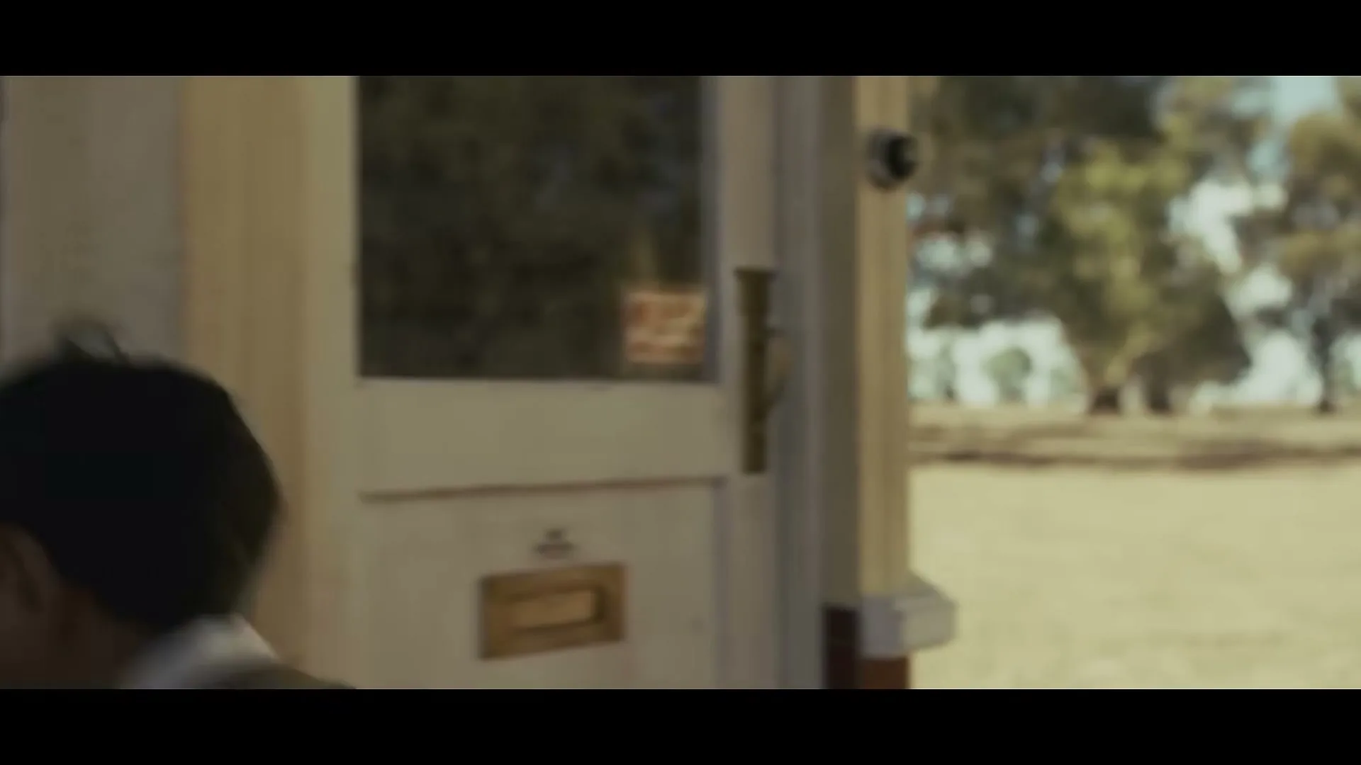 A young boy in a vest walks past a window, revealing an older man working inside a shoe repair shop in a rural setting.
