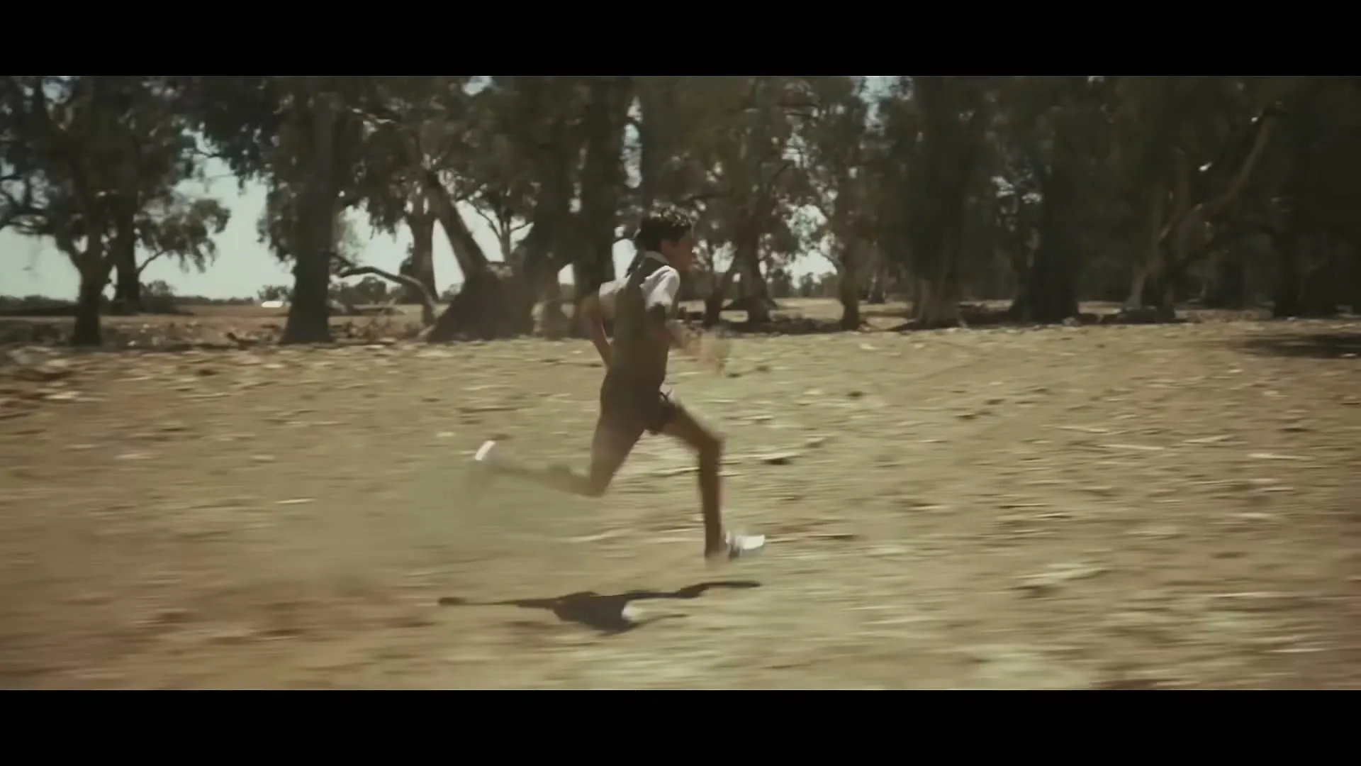A young boy in a vest and shorts runs across a dry, dusty field with trees in the background under a bright sky.